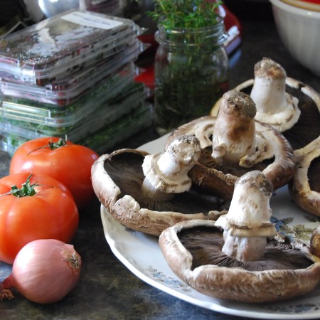 Portabella mushrooms on a plate beside tomatoes and shallots.
